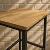 Close-up of a bar stool with a wooden seat and black metal legs against a brick wall. Produktbild-Vorschau