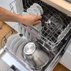 A hand is placing a whisk attachment into a dishwasher filled with dishes and a large metal bowl. Produktbild-Vorschau