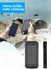 A person is holding a smartphone and a solar power bank outdoors near water and rocks. Produktbild-Vorschau