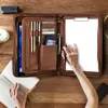 A brown leather portfolio organizer on a wooden desk with a person's hands interacting with it. Náhľad obrázku produktu