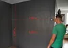 A man is using a laser distance measurer on a dark gray wall, with red measurement lines and numbers overlaid on the image. Produktbild-Vorschau