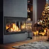 A modern fireplace with a roaring fire is the focal point of a cozy living room decorated for Christmas, with a lit tree and candles in the background. Produktbild-Vorschau