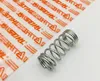 A metal spring is placed on a white surface next to a piece of transparent plastic packaging with orange text. Produktbild-Vorschau