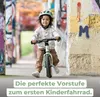 A young child wearing a helmet rides a balance bike in an urban setting with graffiti-covered walls. Produktbild-Vorschau