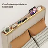 A close-up view of a modern upholstered bed headboard with integrated shelving and lighting, displaying various electronic devices and accessories. Produktbild-Vorschau