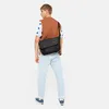 A man is shown from the back wearing a brown vest over a white and blue patterned shirt, light blue jeans, and blue sneakers. He is carrying a black messenger bag across his body. Náhled obrázku produktu