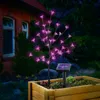 A pink LED blossom tree with a solar panel is planted in a wooden planter box next to a brick house at night. Produktbild-Vorschau