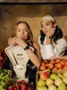 Two young women are posing in a market stall setting, surrounded by fruits and a vintage scale. Voir les images du produit