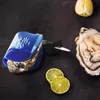 An oyster knife with a blue silicone holder, an opened oyster, lime slices, and a lemon wedge on a dark, textured surface. Produktbild-Vorschau