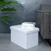 A white storage ottoman sits on a tiled floor in front of a dark gray tiled wall and a portion of a gray sofa. Two books are stacked on top of the ottoman. Náhled obrázku produktu