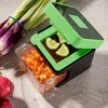 A vegetable chopper with chopped vegetables in a container and some raw vegetables nearby. Produktbild-Vorschau