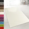 A cream-colored shaggy rug is laid out on a light-colored wooden floor in a room with white curtains and a white nightstand. Náhled obrázku produktu