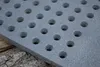A close-up of a gray metal grate with circular holes, resting on a wooden surface. Voir les images du produit