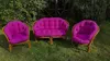 Three pieces of rattan furniture with fuchsia cushions are placed on a grassy lawn in a garden setting. Produktbild-Vorschau