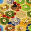 A close-up view of a board game, likely Settlers of Catan, showing hexagonal tiles with numbers and resource illustrations, along with game pieces. Náhľad obrázku produktu