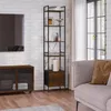 A tall, narrow bookshelf with a rustic brown finish and black metal frame is placed in a living room setting next to a sofa and a media console. Produktbild-Vorschau