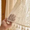 A hand holding a cream-colored lace curtain with tassels. Produktbild-Vorschau