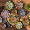 A collection of colorful, patterned ceramic plates and bowls arranged on a wooden table with a brown cloth backdrop. Náhled obrázku produktu