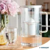 A glass water filter pitcher and a glass of water are placed on a wooden surface next to a vase of pink peonies. Podgląd zdjęcia produktu