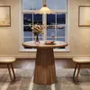 A round wooden dining table with a vase of flowers and a plate of food is set up in front of a large window overlooking a city at dusk. Two stools are placed on either side of the table. Produktbild-Vorschau