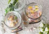Two glass jars filled with wrapped candies are displayed on a patterned tablecloth, with greenery and white roses in the background. Produktbild-Vorschau