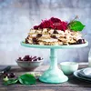 A cake decorated with flowers and cherries on a cake stand, with a bowl of cherries and other items on a wooden table. Produktbild-Vorschau