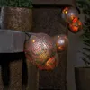 Solar powered string lights with decorative globes are displayed outdoors, possibly on a patio or garden feature, with plants in the background. Produktbild-Vorschau