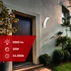 An outdoor wall lamp is mounted on a textured wall next to a dark wooden door. The lamp is white and has a curved design. There are plants and bushes around the entrance. A red overlay in the bottom left corner displays product specifications. Náhled obrázku produktu