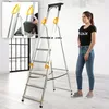 A woman stands next to a silver and yellow aluminum ladder with 6 steps in a room with large windows. Náhled obrázku produktu