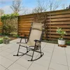 A beige rocking chair is placed on a tiled patio next to a wooden fence and some plants. Produktbild-Vorschau
