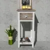 A white and brown nightstand with a drawer and a shelf, placed against a textured gray wall. A vase of white tulips is on the left, and a book is on the shelf. Podgląd zdjęcia produktu
