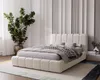 A modern bedroom with a large upholstered bed as the focal point, decorated with pillows and a throw blanket. A large potted plant is to the left, and a textured wall is in the background.