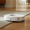 A white robot vacuum cleaner is shown on a rug and hardwood floor, transitioning between the two surfaces. Náhled obrázku produktu