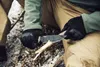 A person wearing tactical gloves and clothing is using a knife to shave wood in an outdoor, possibly snowy environment. Produktbild-Vorschau