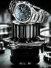 A close-up shot of a silver watch with a black face, resting on a metal gear and other mechanical parts. Produktbild-Vorschau