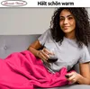 A woman is relaxing on a couch, wrapped in a pink blanket and holding a glass of red wine and a remote control. Produktbild-Vorschau