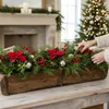 A person is decorating a wooden planter box with Christmas greenery, poinsettias, berries, pinecones, and ornaments. A fireplace and Christmas tree are visible in the background. Podgląd zdjęcia produktu