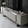 A modern TV stand with a travertine-like finish and silver accents, placed in a dimly lit room with a large black television above it. Voir les images du produit