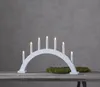 A white arch candle holder with seven electric candles sits on a wooden table in front of a gray wall. Two small mounds of green moss are placed on either side of the candle holder. Produktbild-Vorschau