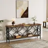 A long, narrow console table with a rustic wood top and a black metal base with a decorative pattern is shown in a well-lit room. The table is adorned with a lamp, a bowl of nuts, a vase with greenery, and decorative boxes. A framed abstract painting hangs Náhľad obrázku produktu