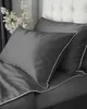 A close-up view of dark gray bedding with white piping, set against a tufted headboard and decorative branches. Náhled obrázku produktu