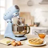 A pastel blue SMEG stand mixer is shown on a kitchen counter, with ingredients for baking like butter, eggs, and a pie, as well as a bowl of jam. Anteprima dell’immagine prodotto