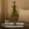 A small, artificial Christmas tree with LED lights sits on a table next to a decorative pot. The background is a softly lit room with curtains and a pillow. Produktbild-Vorschau