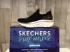 A black Skechers D'Lux Walker sneaker is displayed on top of its packaging box, with a wood-paneled background. Náhled obrázku produktu