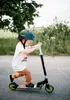 A young boy wearing a helmet rides a scooter outdoors on a paved path. Produktbild-Vorschau