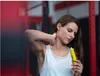 A woman is holding a yellow tube and applying cream to her neck in a gym setting. Produktbild-Vorschau