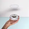 A hand holding a smoke detector with batteries inserted, with the mounting plate visible on the ceiling above. Produktbild-Vorschau