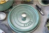 A close-up view of a green Staub pot lid with a silver handle, placed on a gray surface. Other kitchen items like a salt dish and a corkscrew are visible. Produktbild-Vorschau