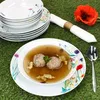 A table setting with a soup bowl, stacked plates, a napkin, and a poppy flower on a green grassy surface. Produktbild-Vorschau