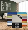 A stack of folded fitted sheets in various colors, with a logo and text overlay on the front sheet. The background shows a bedroom setting with a bed and a wall with a large graphic logo. Produktbild-Vorschau